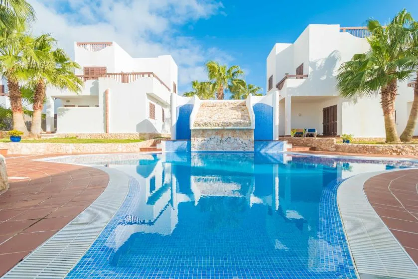 Una piscina está entre edificios blancos y palmeras bajo un cielo azul.