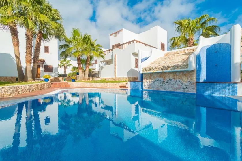 Una piscina azul refleja edificios blancos y palmeras bajo un cielo despejado.