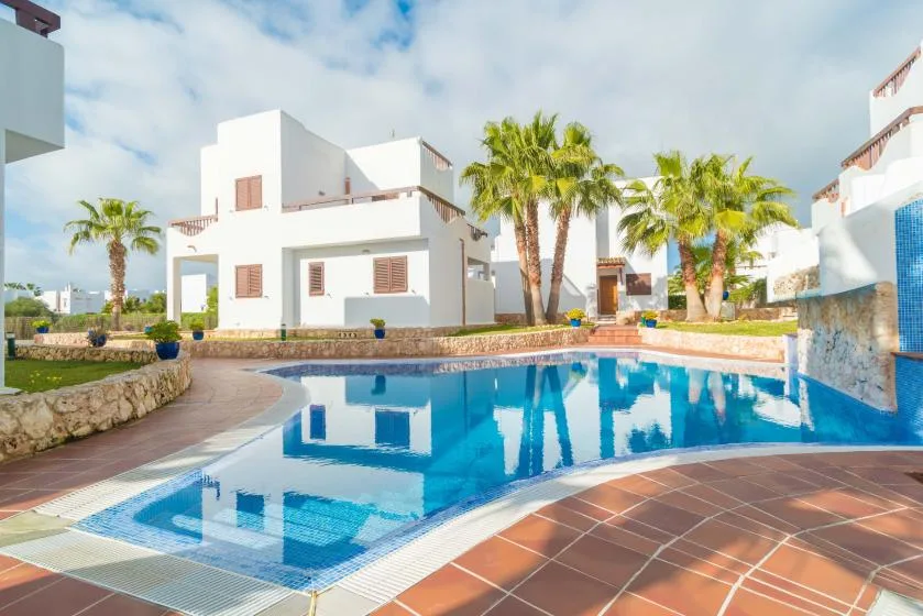A swimming pool sits among white buildings and palm trees in a sunny courtyard.