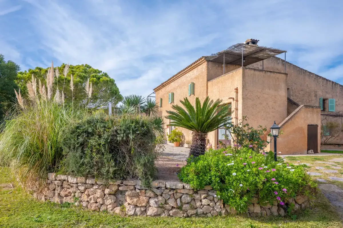 A tan building stands behind a stone garden wall and flowering plants.