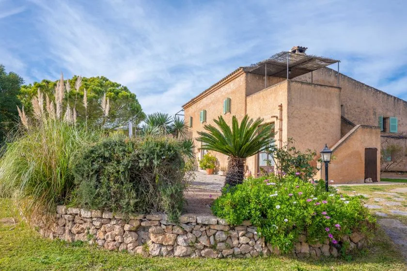 A tan stone building stands behind a landscaped garden with flowering shrubs.