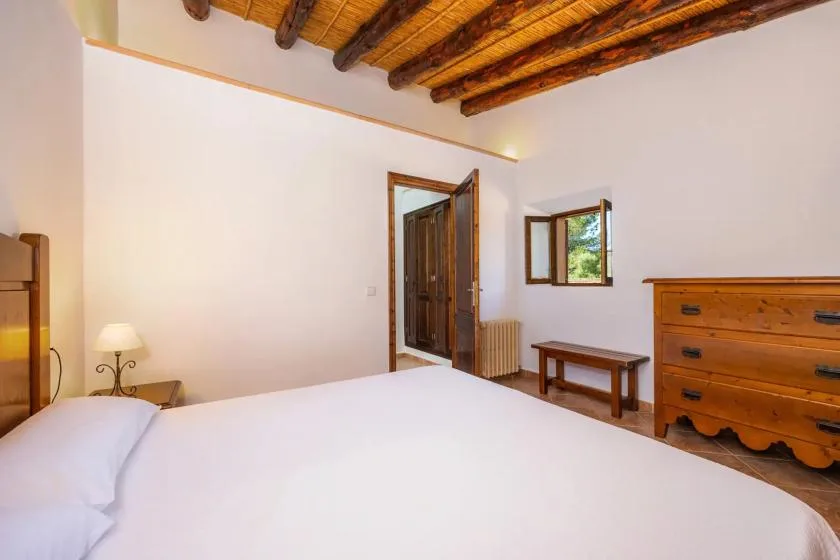 A bedroom with a white bed, wooden ceiling beams, and a window.