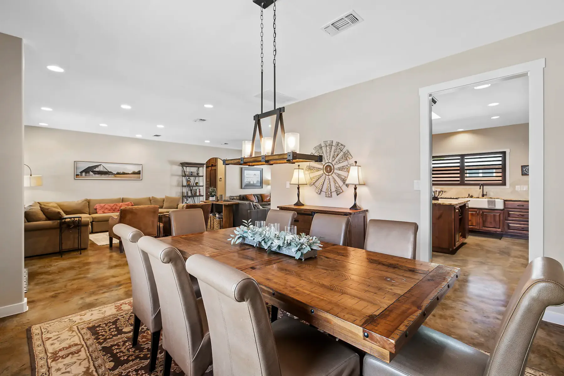 Modern dining area with a wooden table, beige chairs, rustic chandelier, and adjacent living room and kitchen in the background. Neutral tones.