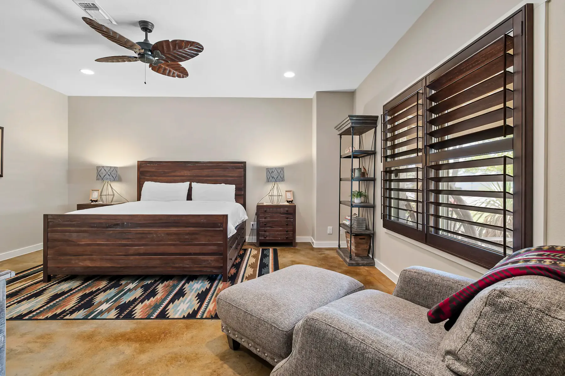 Dormitorio amplio con cama de madera, alfombra estampada, sillón, reposapiés y ventana grande. Los tonos neutros y la decoración ecléctica realzan la calidez.