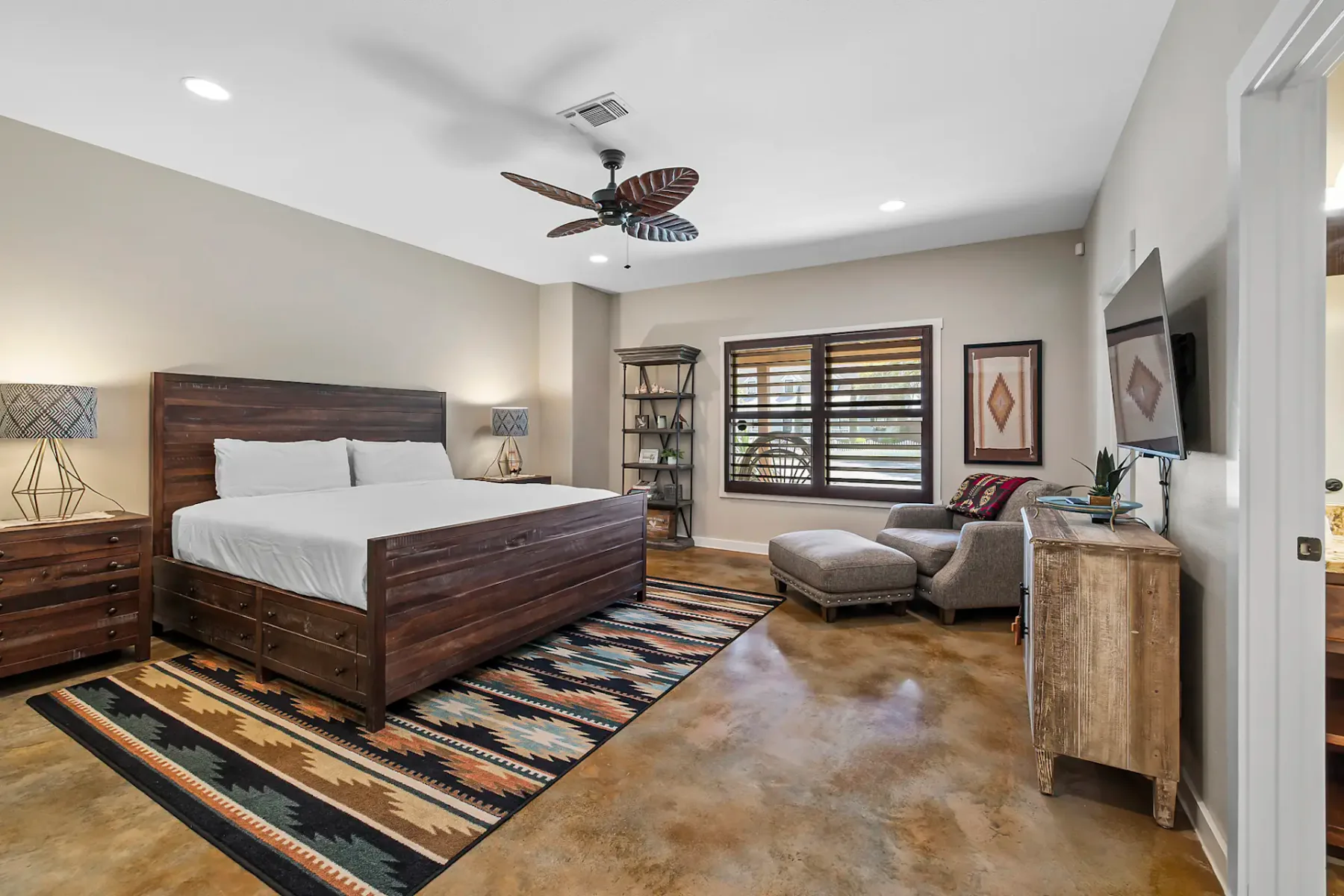 Dormitorio amplio con cama de madera, alfombra estampada, sillón, reposapiés y ventana grande. Los tonos neutros y la decoración ecléctica realzan la calidez.