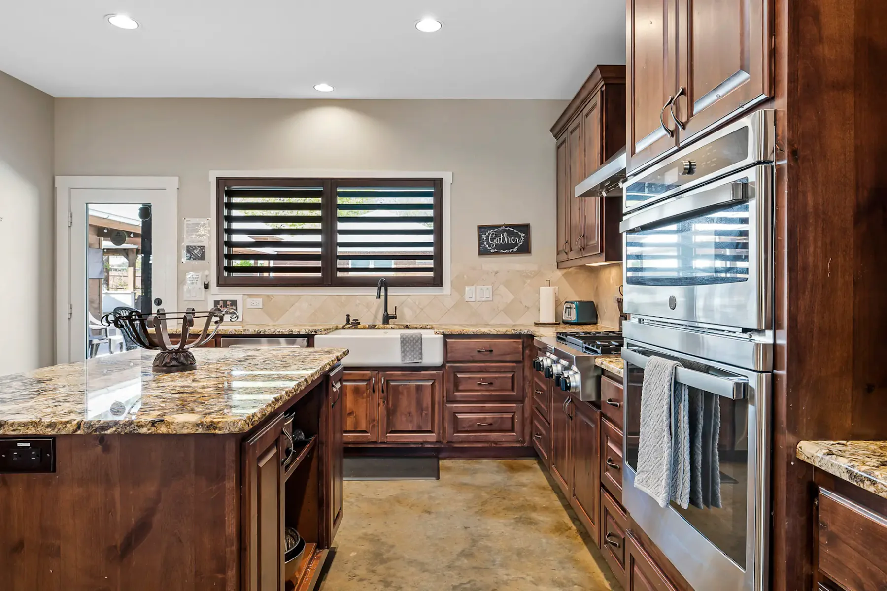 Modern kitchen with granite countertops, wooden cabinets, double ovens, farmhouse sink, and utensil holder on island. Door and window with blinds.