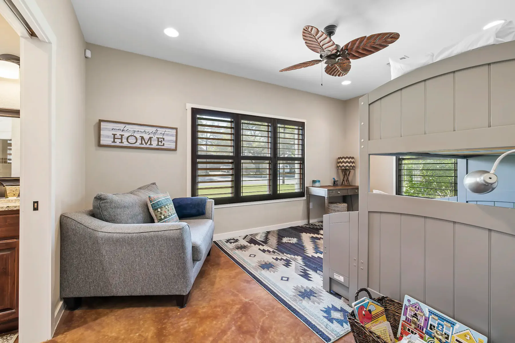 Habitación acogedora con sofá gris, alfombra decorativa, ventana con contraventanas, ventilador de techo y un pequeño escritorio. El cuadro de la pared dice