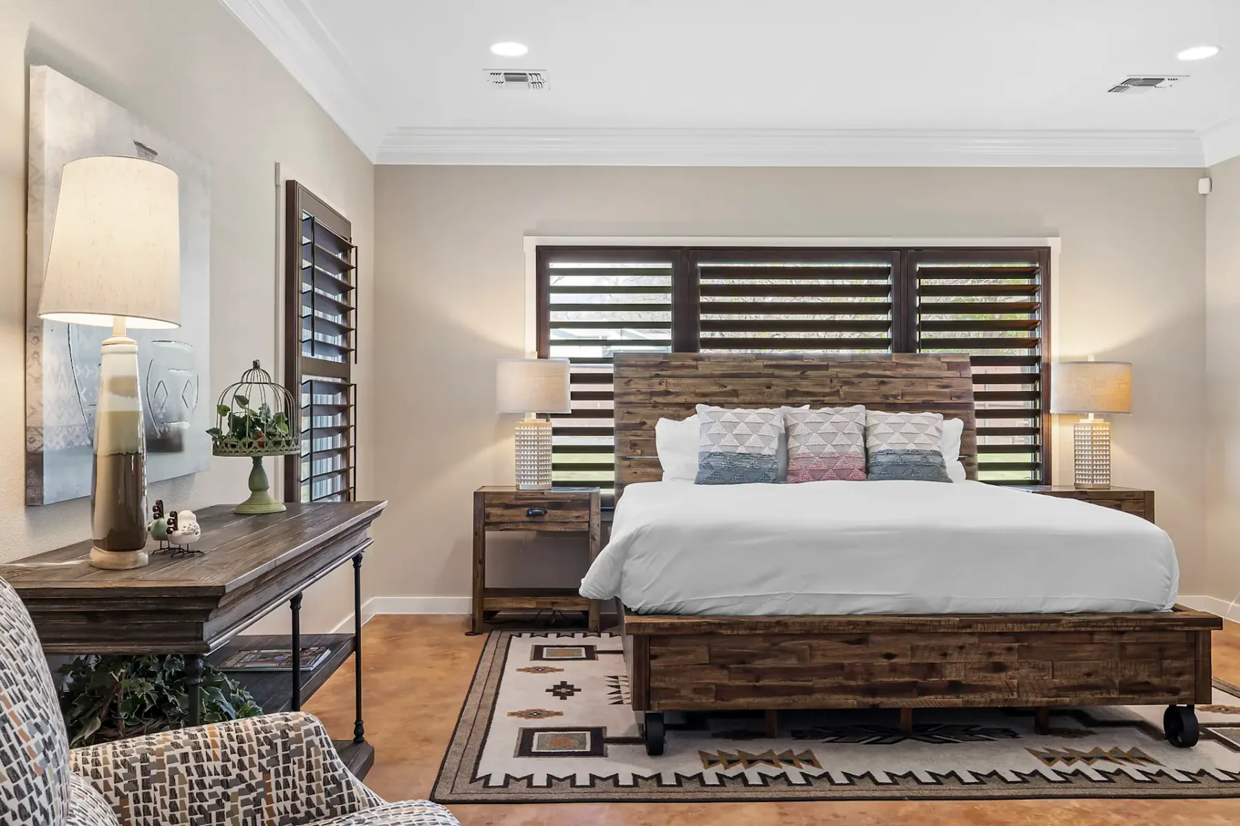 Dormitorio acogedor con cama de madera, ropa de cama blanca, cojines estampados, dos mesas de noche con lámparas y una alfombra estampada sobre un suelo de madera.