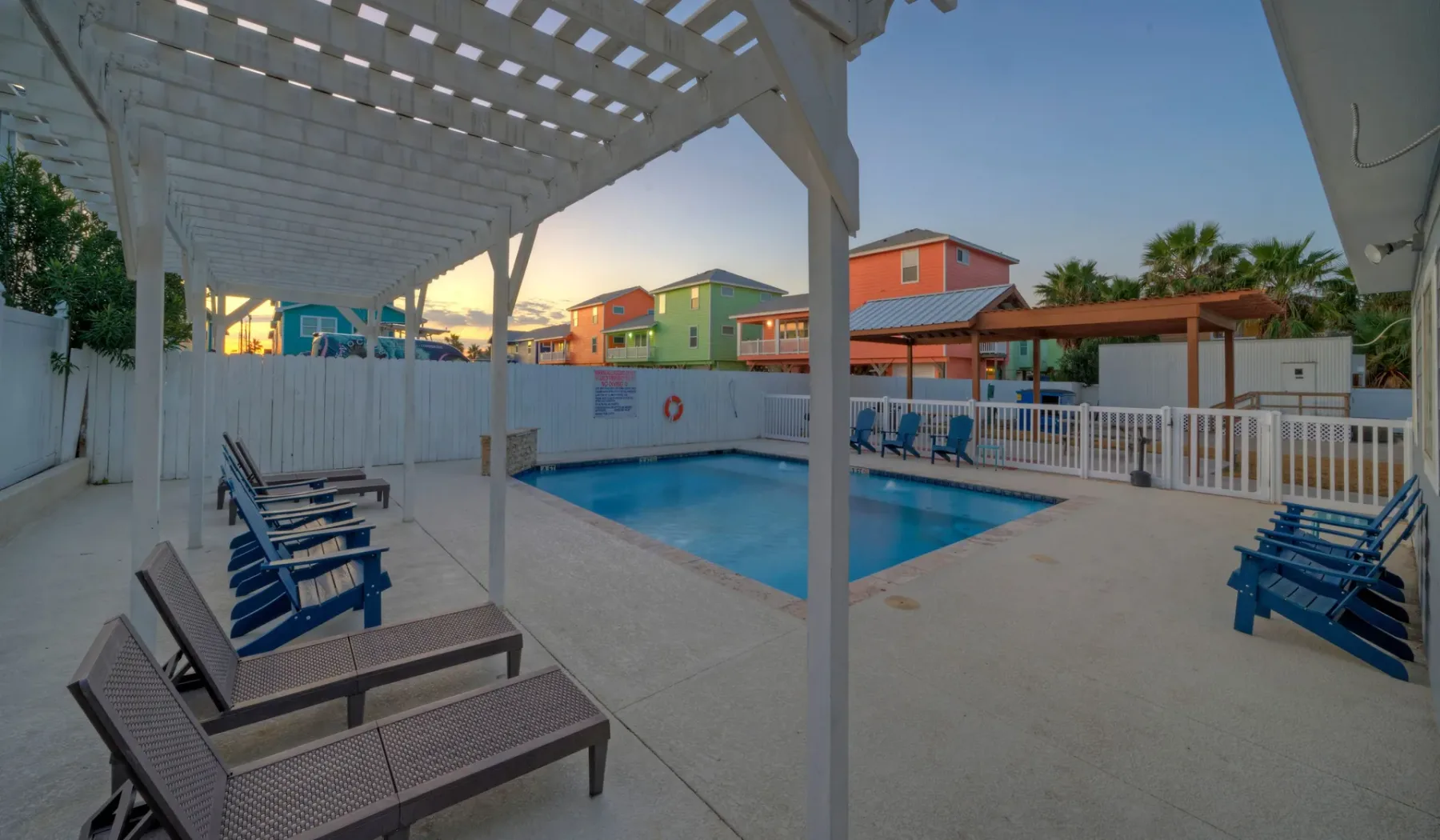 An outdoor pool area has lounge seating beneath a white pergola.