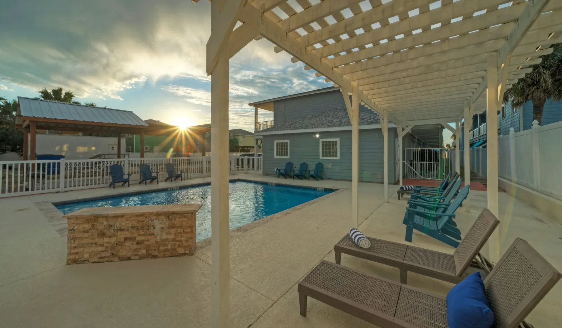An outdoor pool area has lounge seating beneath a wooden pergola.