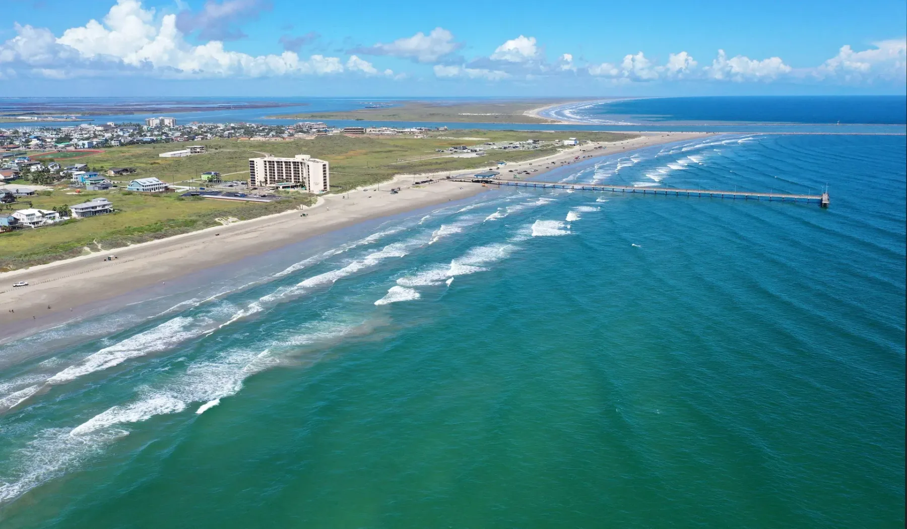 An aerial view of a sandy coastline with waves and buildings along the shore.