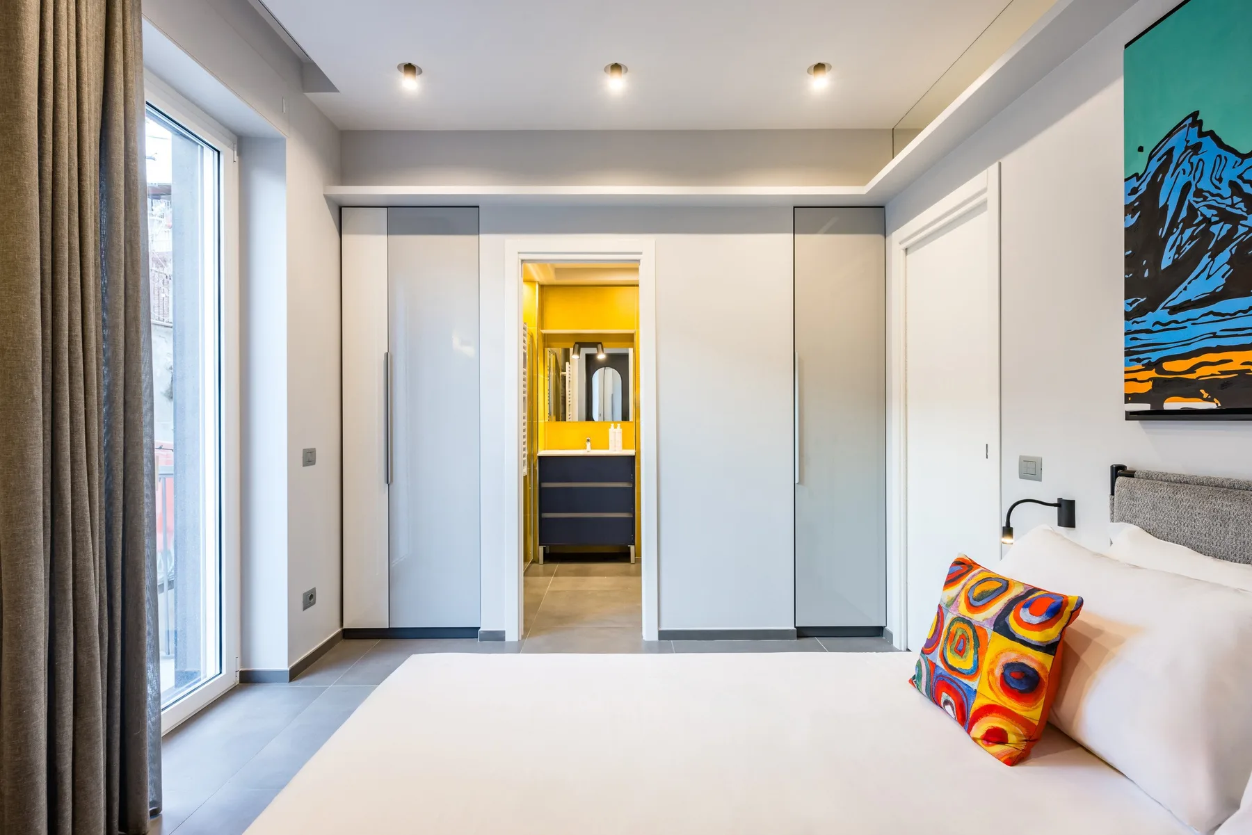 Un dormitorio luminoso con una cama junto a una ventana grande y arte colorido en la pared.