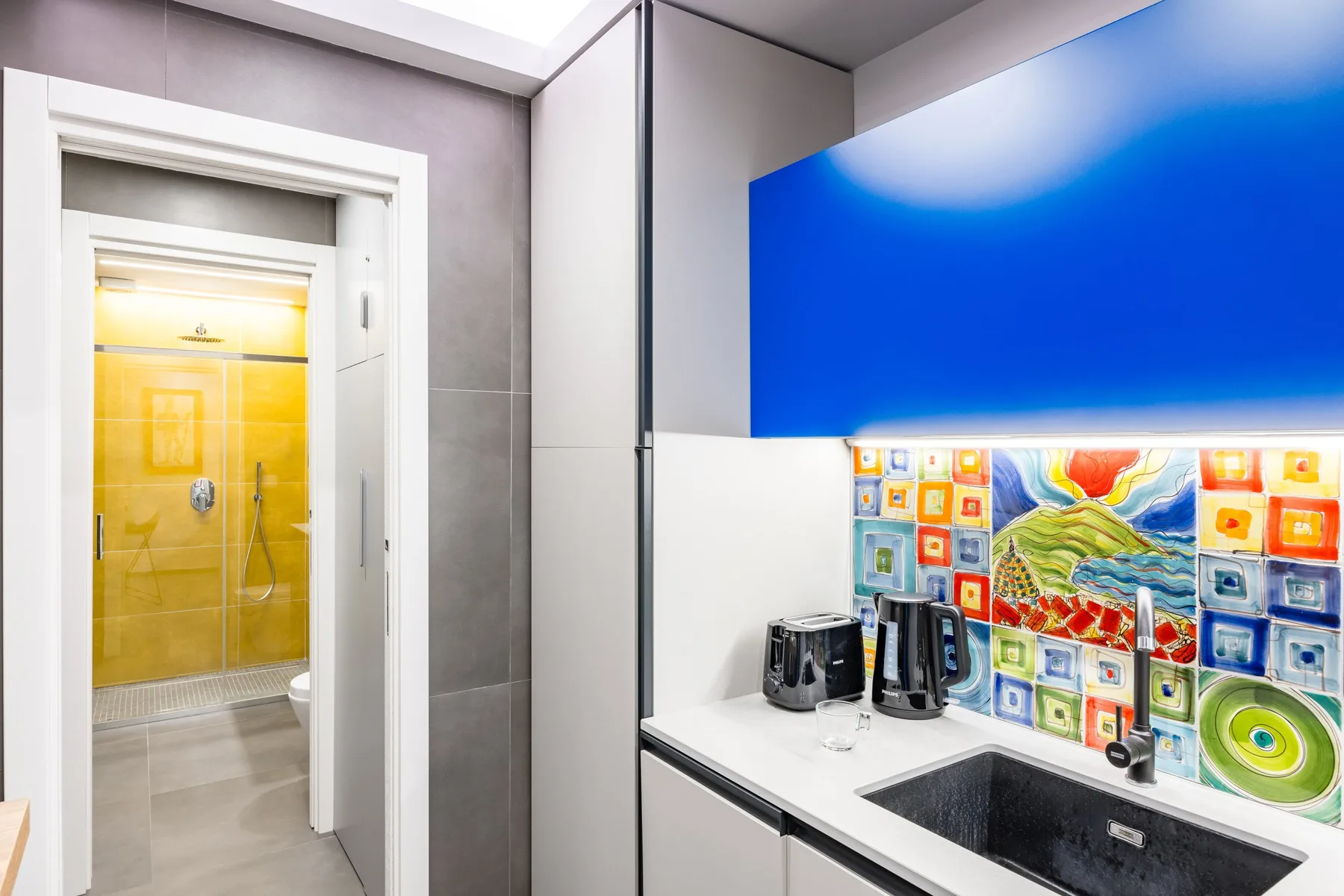 Una pequeña cocina con fregadero oscuro y un salpicadero de azulejos coloridos junto a una puerta de baño.