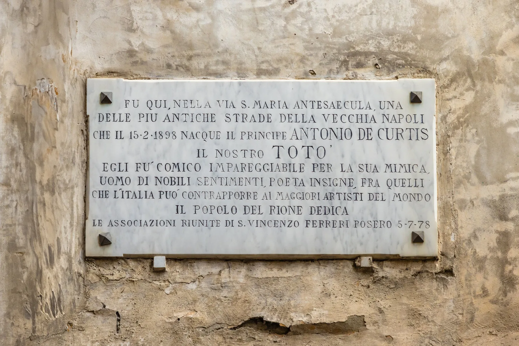 Una placa conmemorativa de piedra con texto italiano grabado está montada en un muro desgastado.