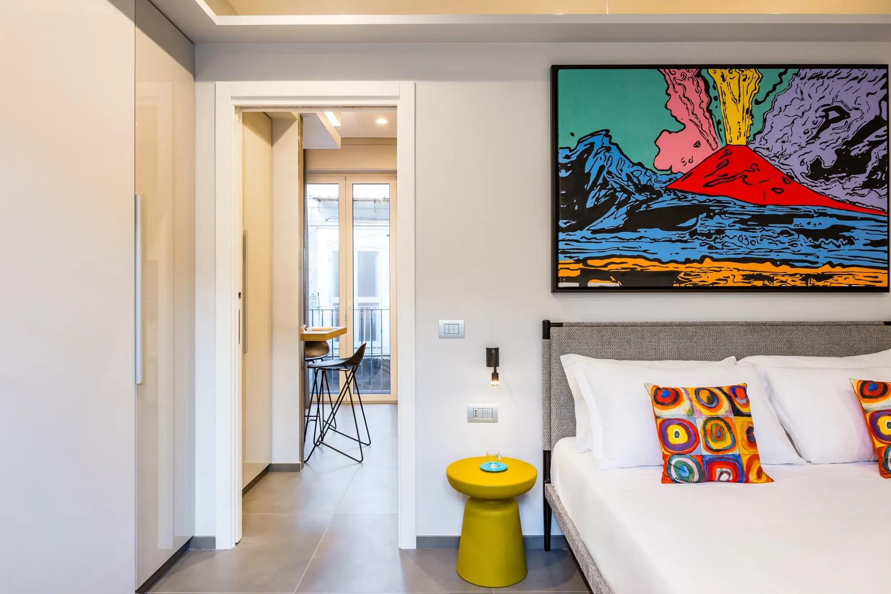Un dormitorio con una cama bajo una obra colorida y una puerta que da a una pequeña zona de comedor.
