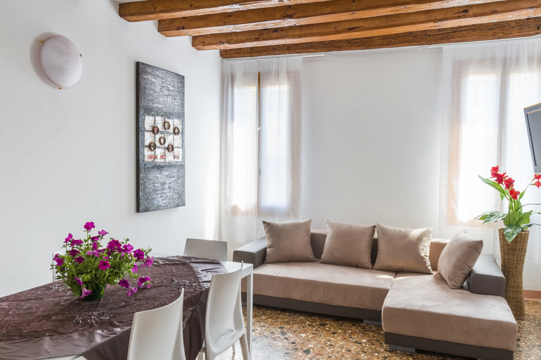 A bright living and dining area has a sectional sofa, dining table, and exposed ceiling beams.