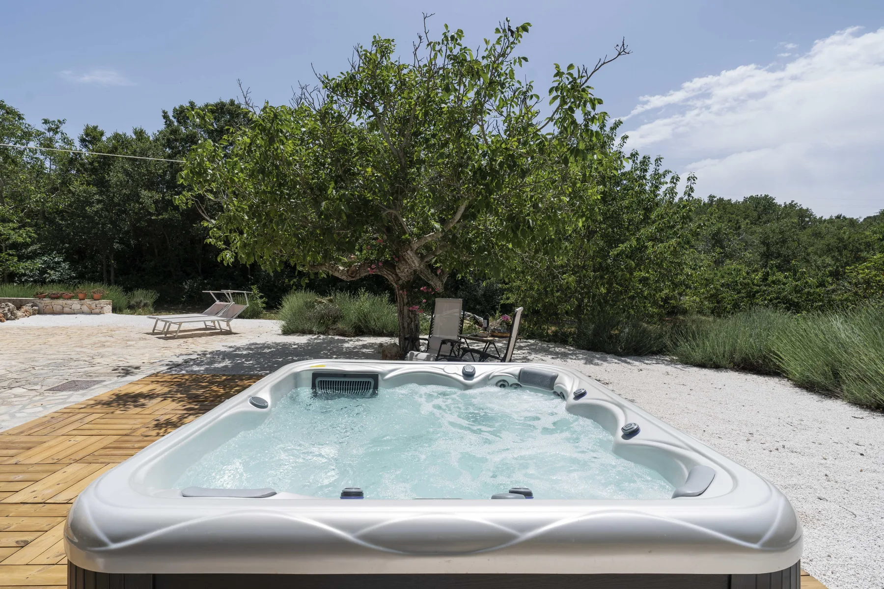 Una bañera de hidromasaje al aire libre está junto a una terraza de madera, bajo un árbol frondoso.