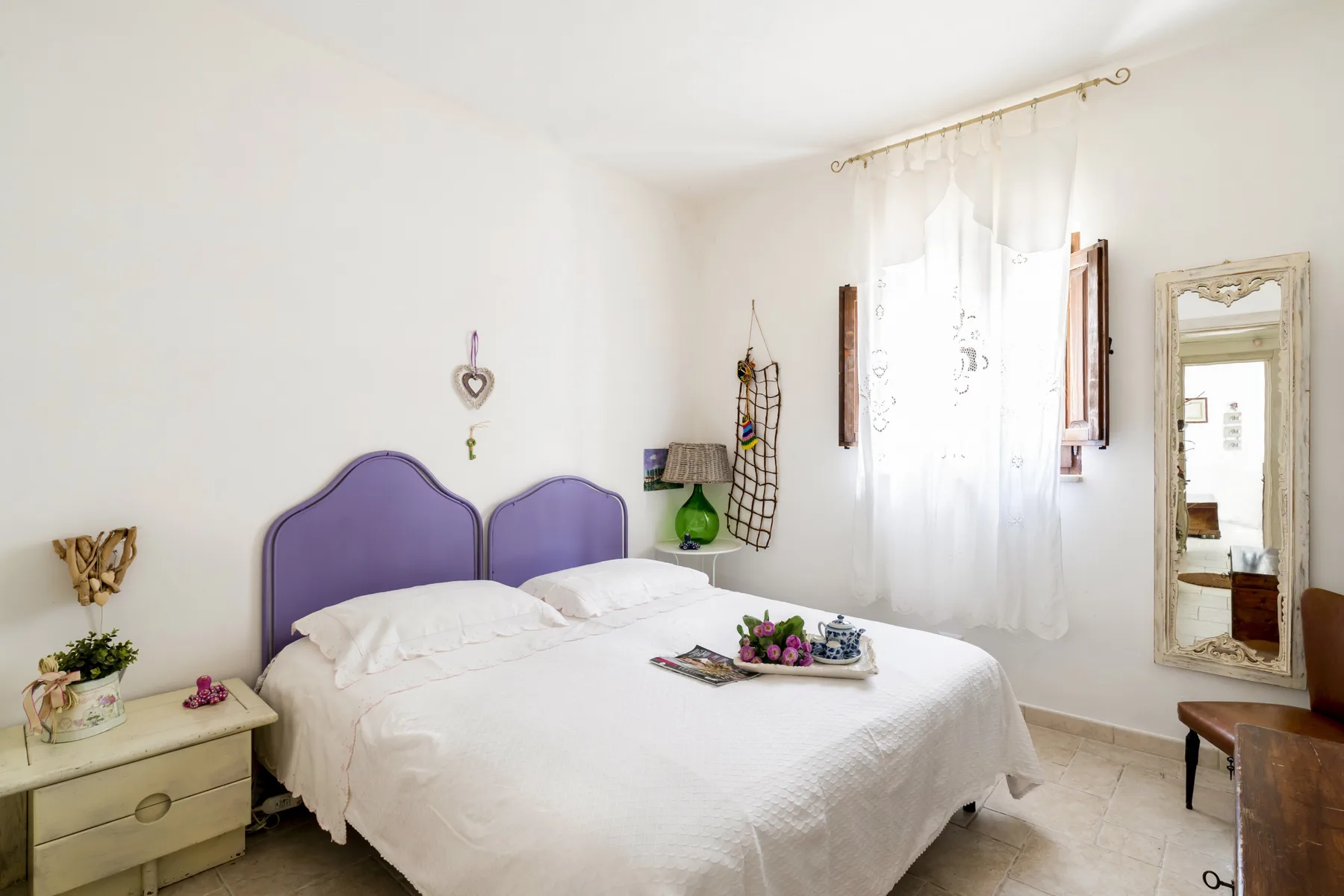 Un dormitorio luminoso con una cama blanca, cabecero morado y cortinas translúcidas.