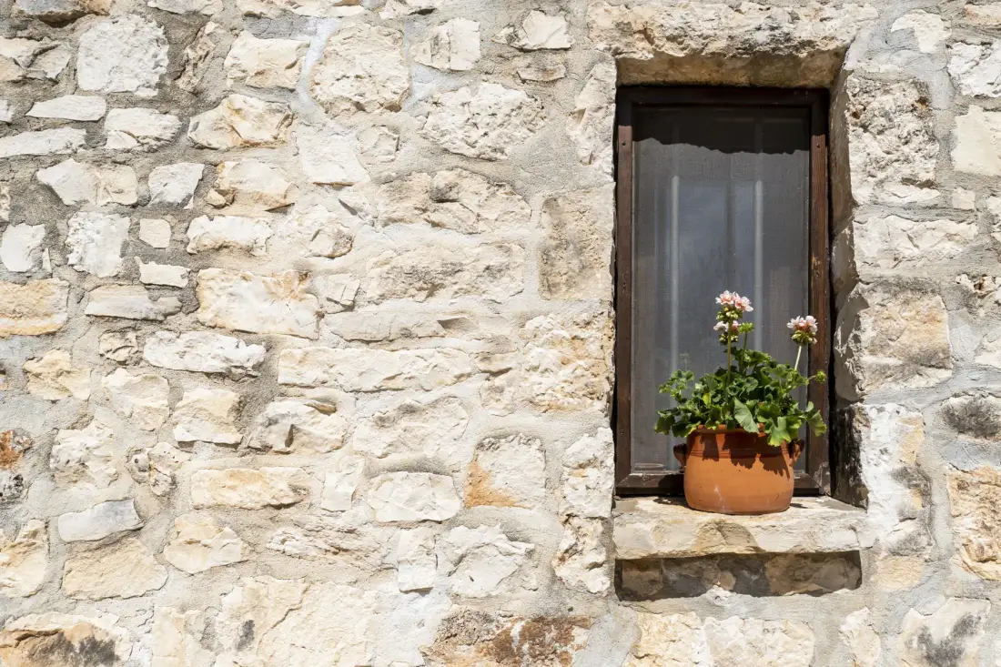 Una ventana pequeña con una planta florida está en un muro de piedra clara.