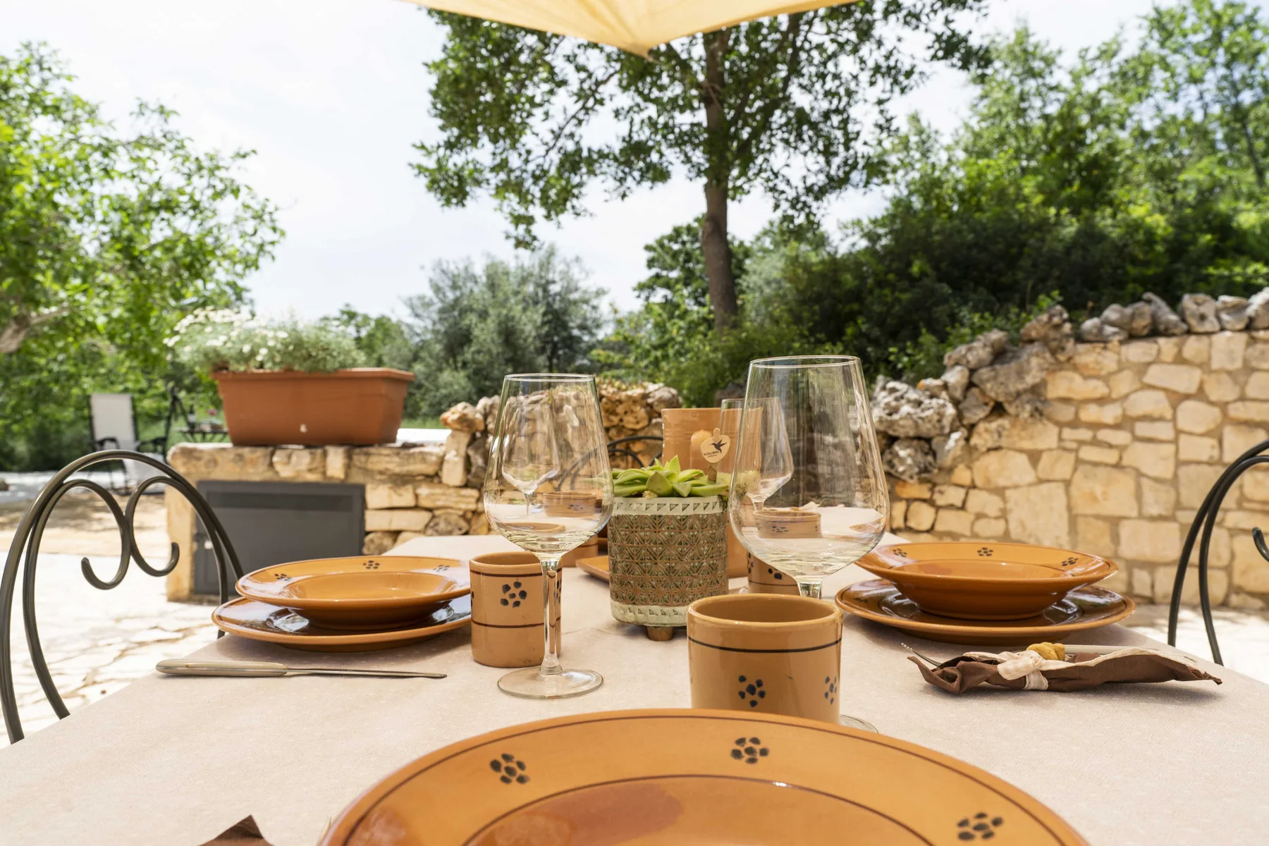 Una mesa de comedor al aire libre está puesta con platos y copas junto a un muro de piedra.