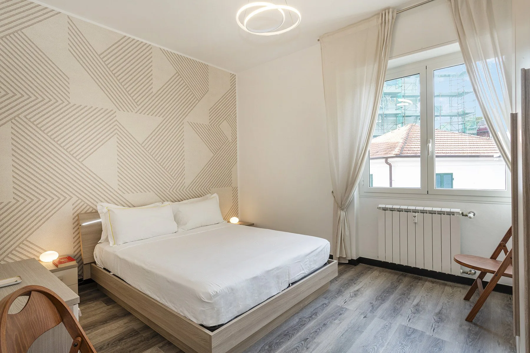 Un dormitorio luminoso con una cama, una pared decorativa estampada y una ventana amplia con cortinas.