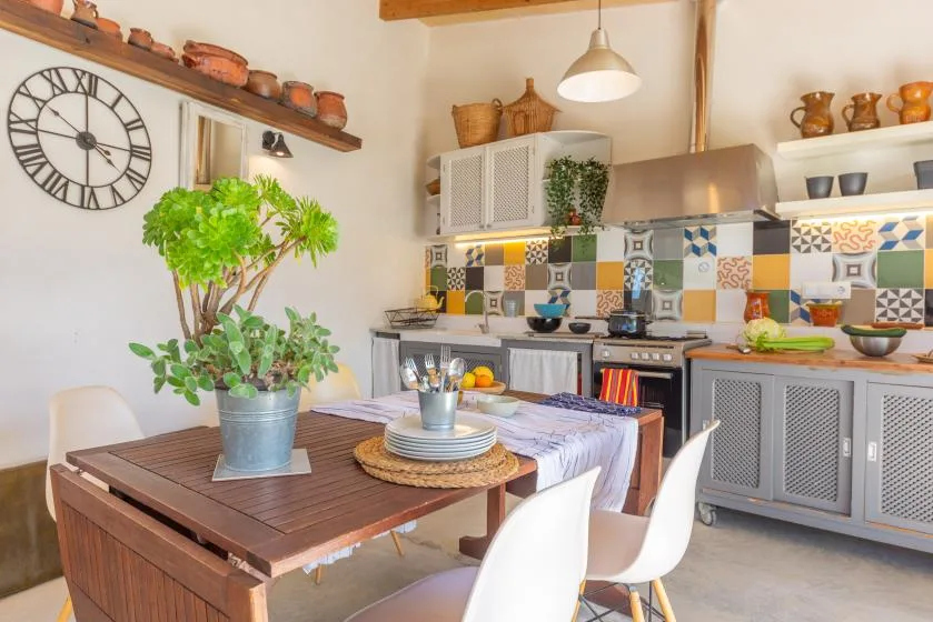 Una mesa de comedor con sillas está en una cocina luminosa con azulejos decorativos de colores.