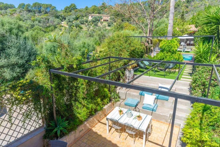 Un patio comedor al aire libre con mesa y sillas bajo una pérgola, rodeado de vegetación.