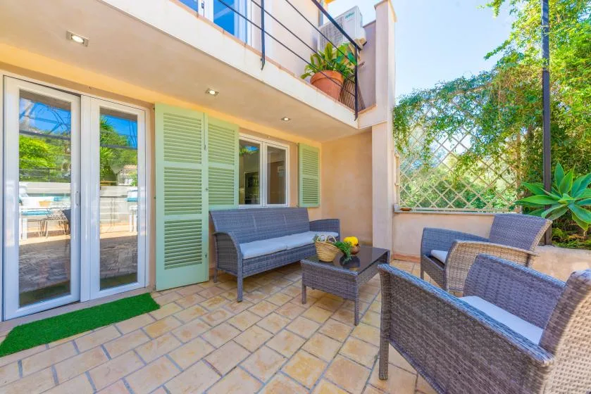 Un patio embaldosado tiene asientos de mimbre junto a puertas de vidrio y persianas verdes.