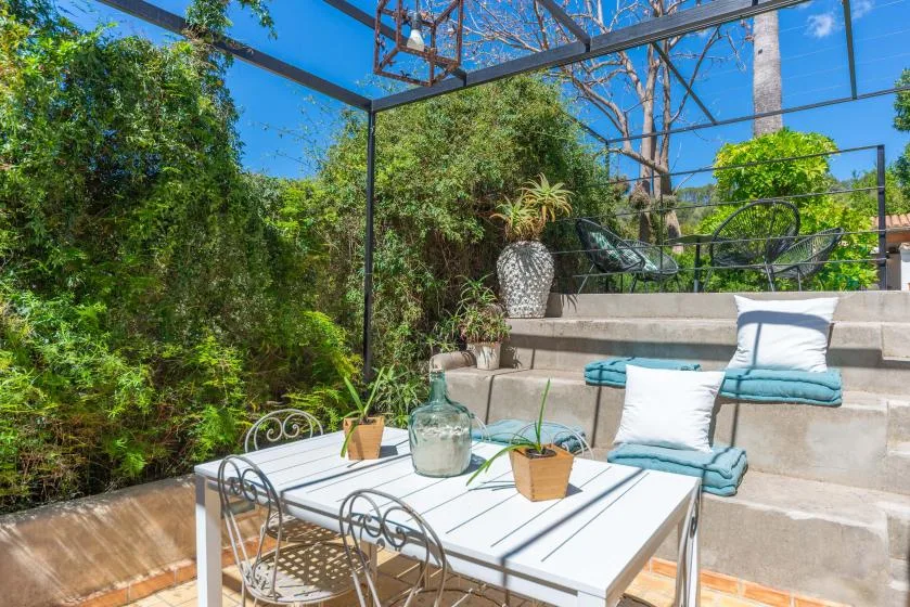 Un patio al aire libre con una mesa blanca, asientos y vegetación alrededor.