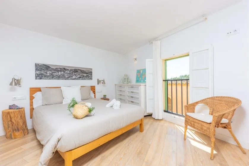Un dormitorio luminoso con una cama y una silla de mimbre junto a una puerta abierta al balcón.