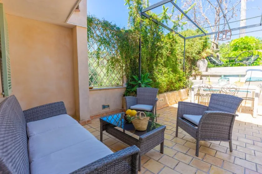 Un patio exterior soleado con asientos acolchados, una mesa de centro y vegetación alrededor.
