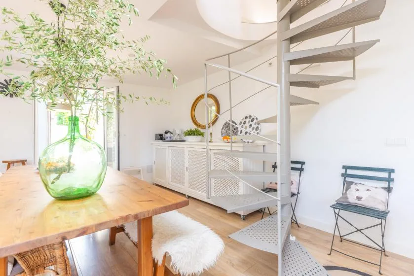 Un comedor luminoso tiene una mesa de madera junto a una escalera de caracol metálica.
