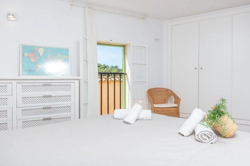 Un dormitorio luminoso con una cama, una silla de mimbre y una puerta abierta al balcón.