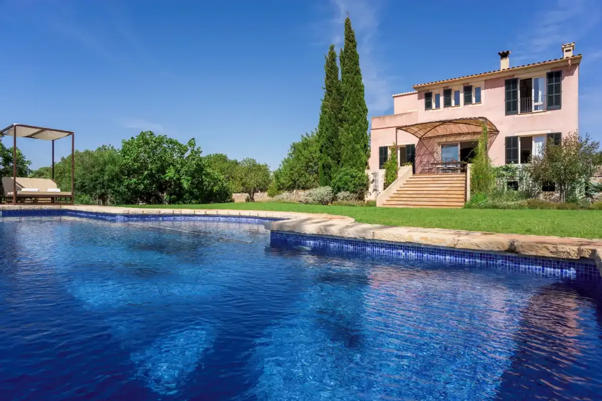 A swimming pool sits in front of a pink two-story house and garden.