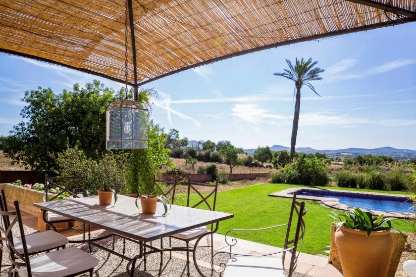 A shaded outdoor dining area overlooks a green lawn, swimming pool, and open landscape.