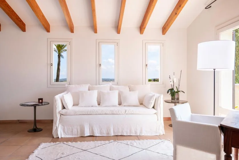 A bright living room with white seating, tall windows, and exposed ceiling beams.