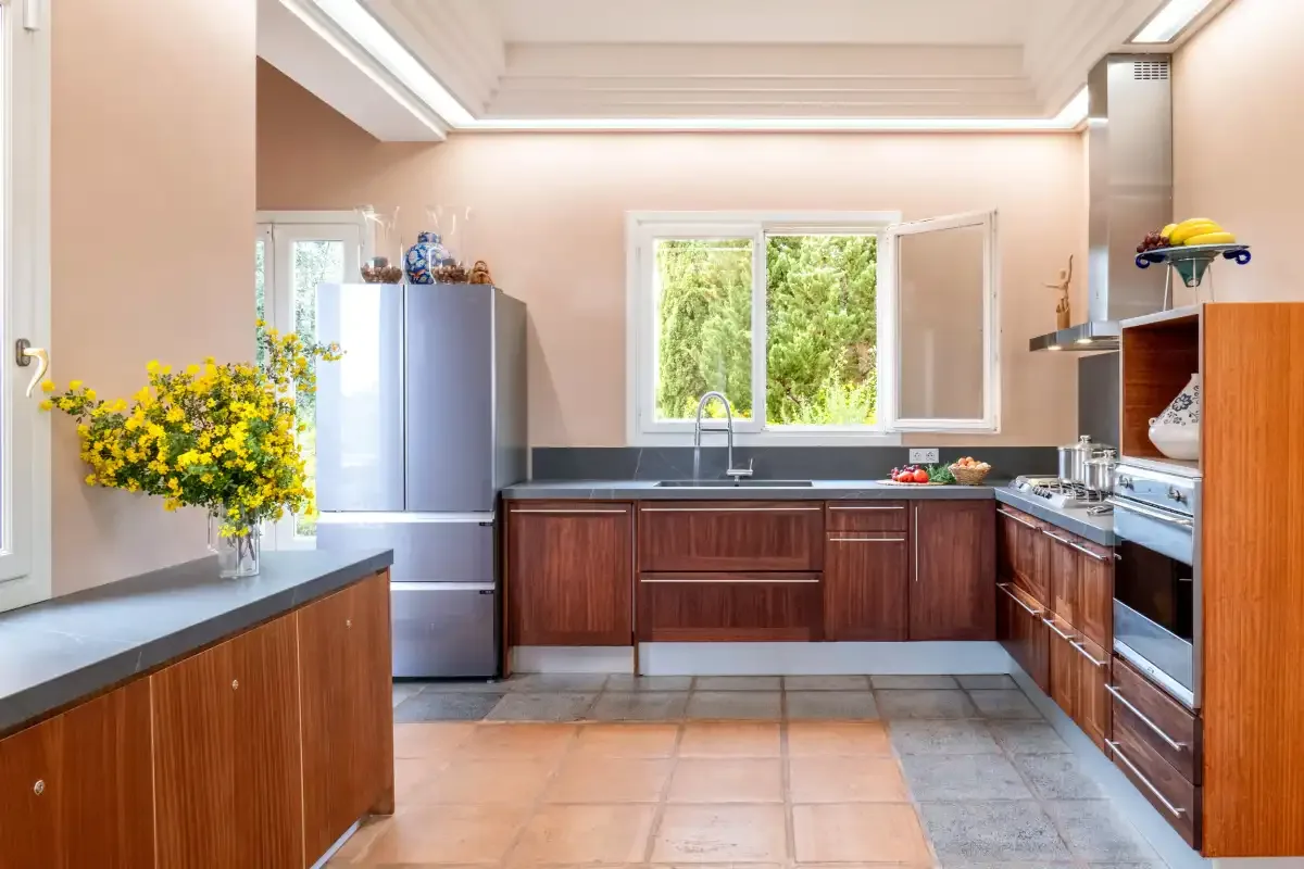A bright kitchen with wooden cabinets, dark countertops, and a window overlooking greenery.