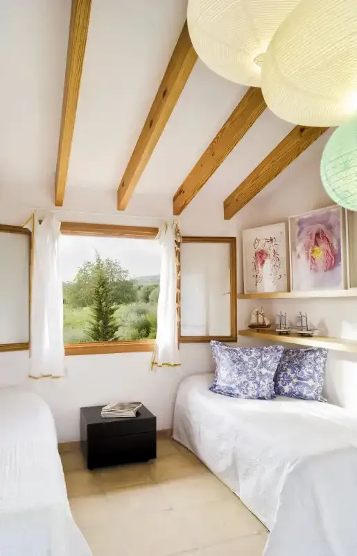 A bright bedroom with white bedding, exposed wooden ceiling beams, and a window overlooking greenery.