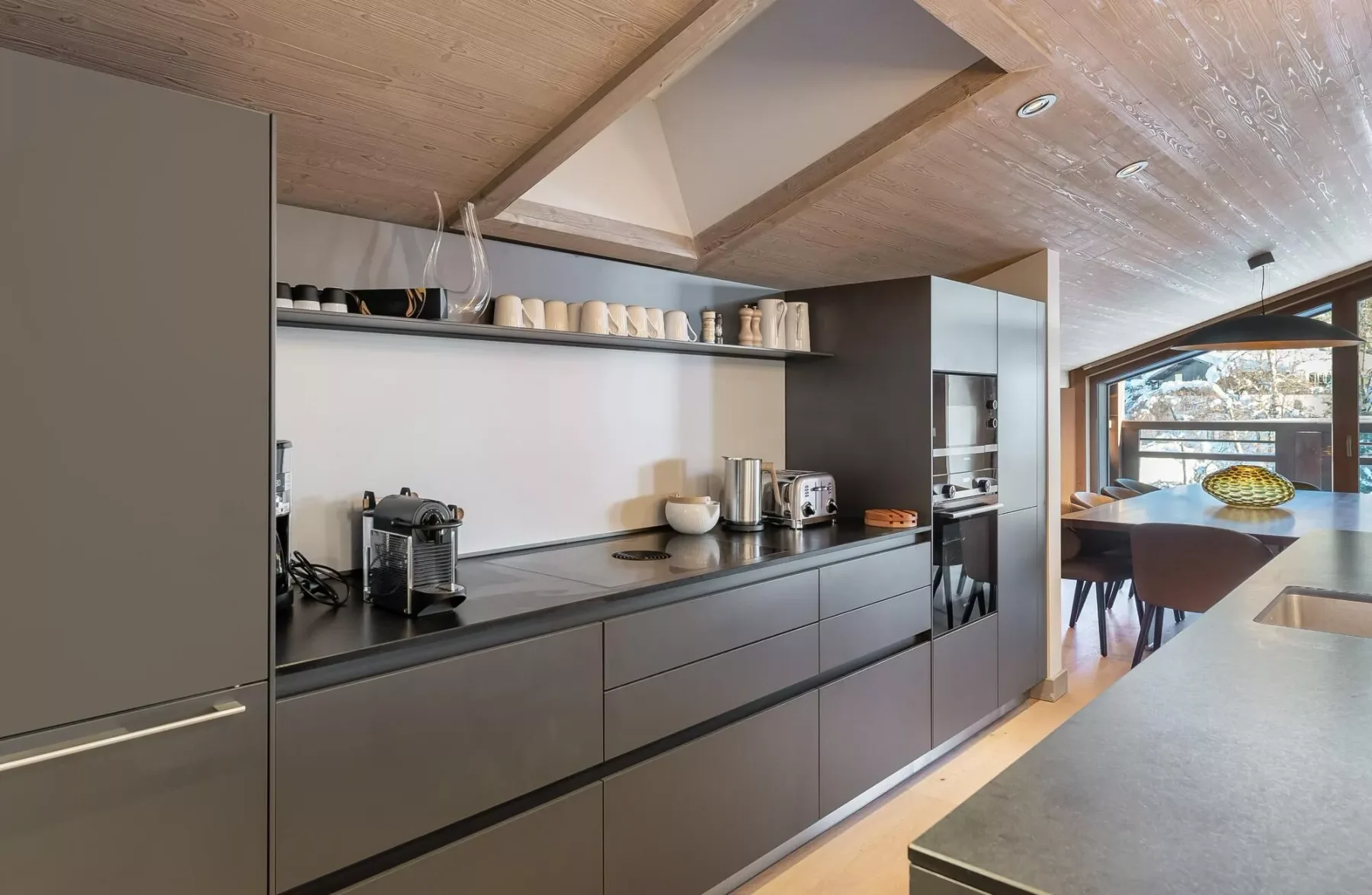 A modern kitchen with dark cabinetry, countertop appliances, and a dining area by the window.