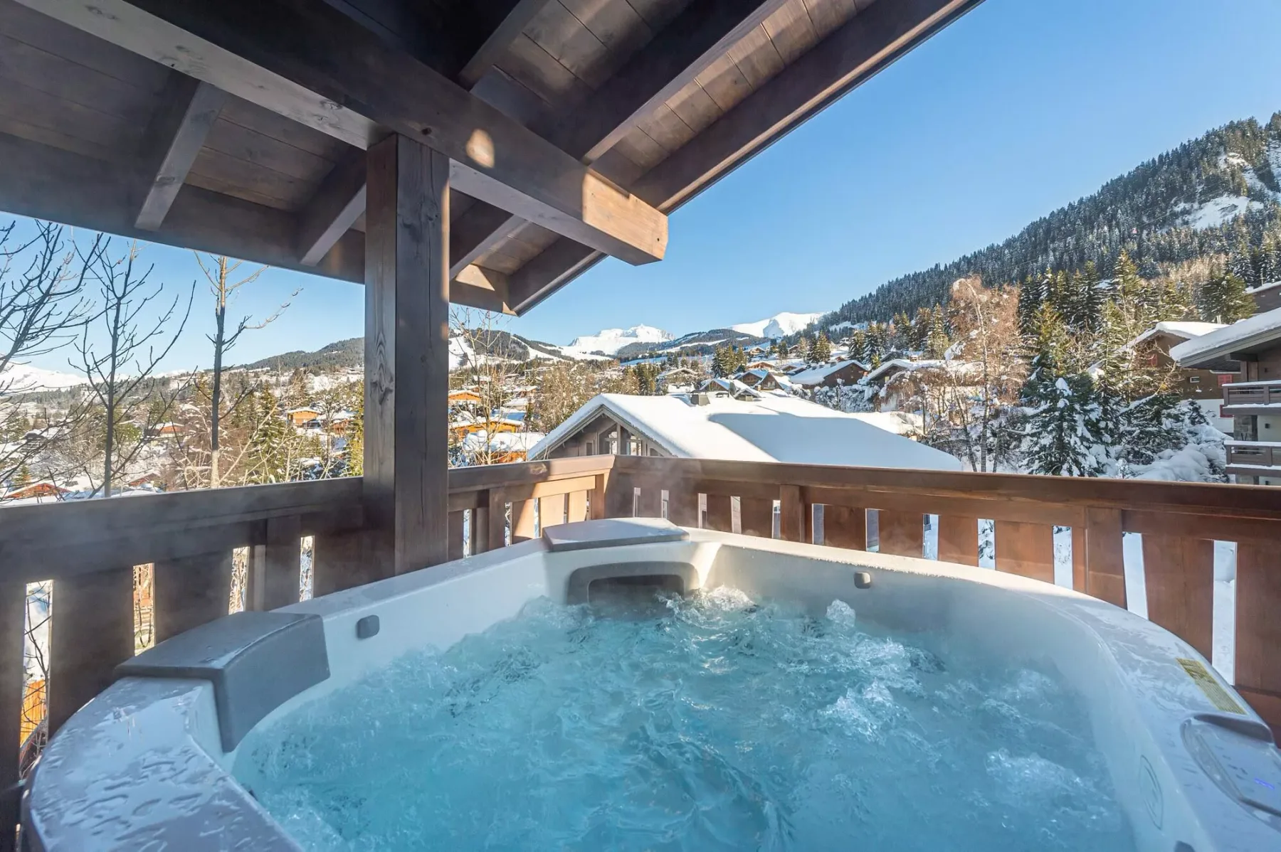 An outdoor hot tub sits on a covered deck overlooking a snowy mountain village.