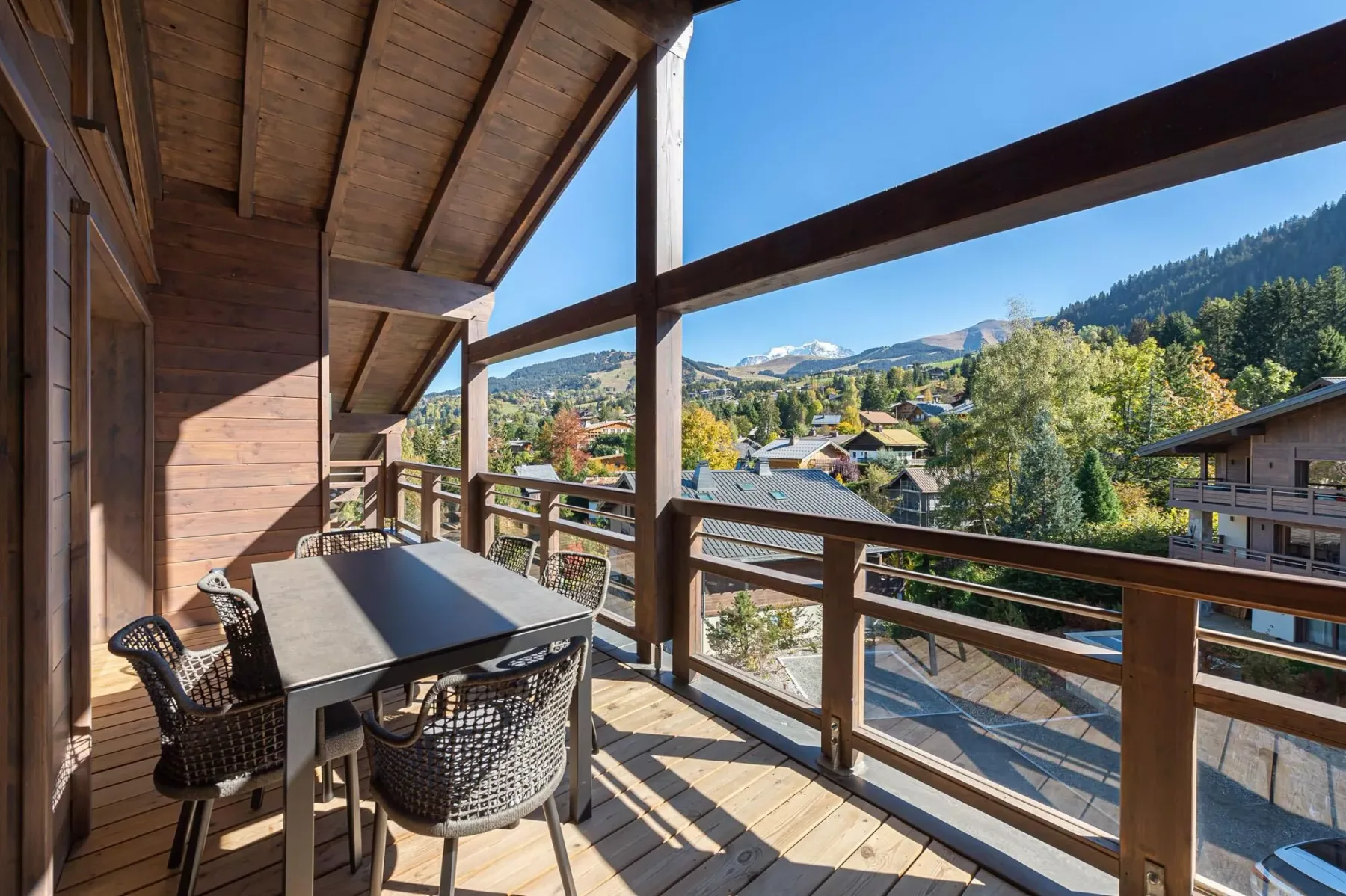 A wooden balcony with outdoor seating overlooks a village and mountain scenery.