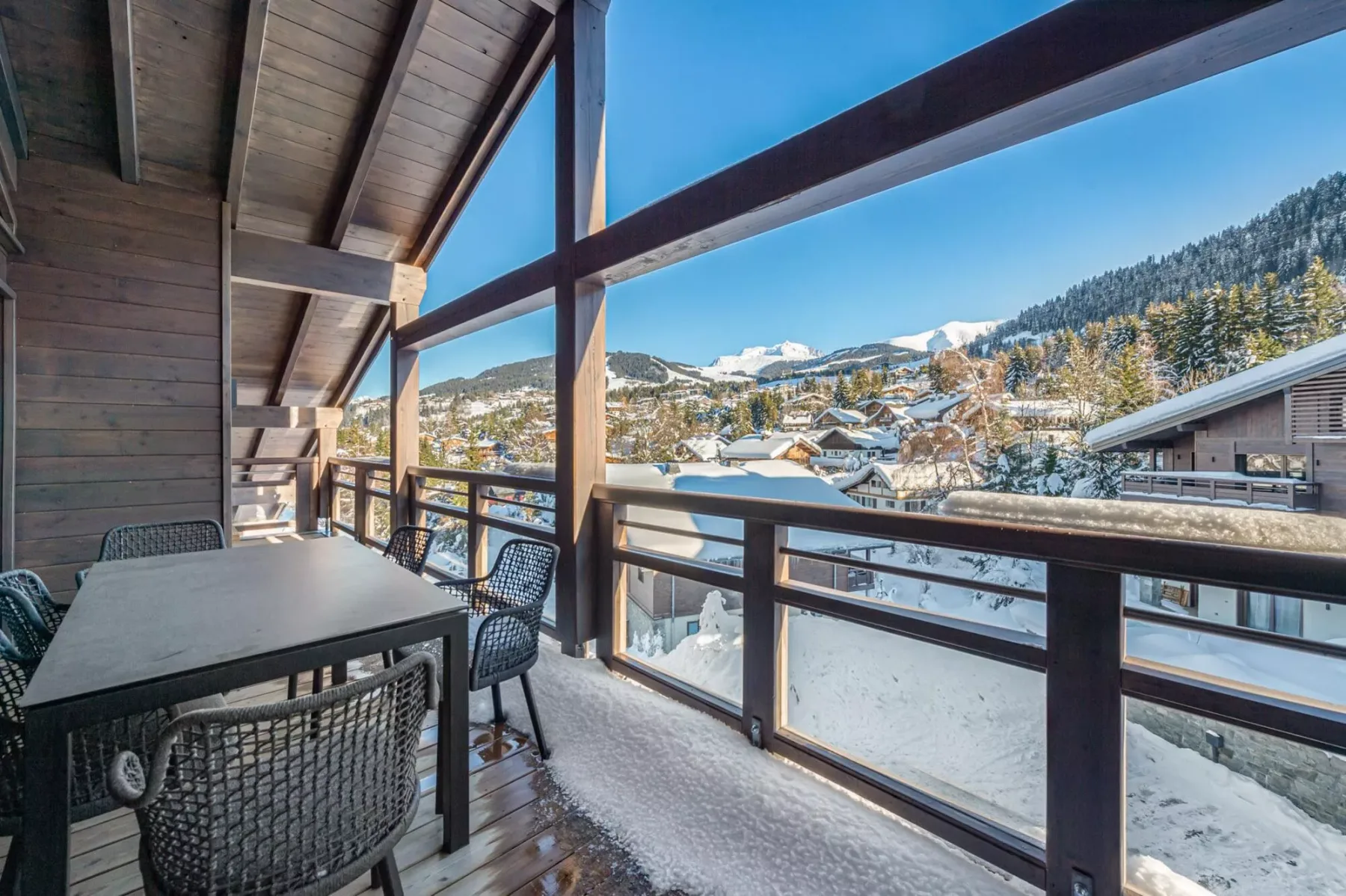 A covered balcony with outdoor seating overlooks a snowy mountain village.