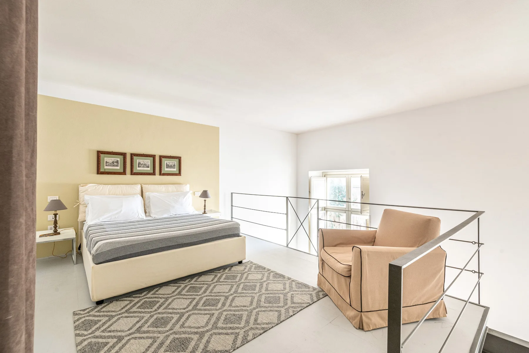 Un dormitorio luminoso tiene una cama, una alfombra estampada y asientos junto a una barandilla.