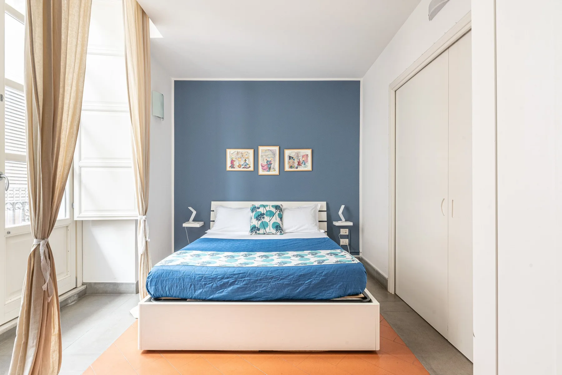 Un dormitorio con una cama frente a una pared azul y ventanas altas.