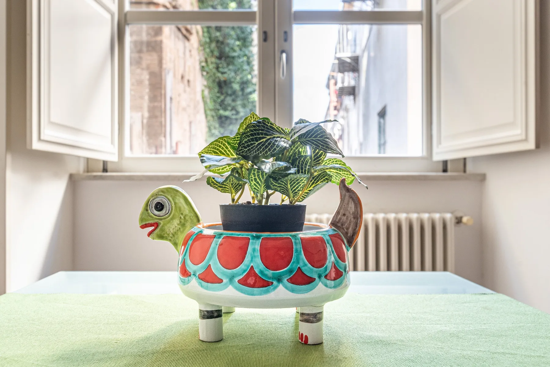 Una maceta colorida con forma de tortuga y una planta frondosa está sobre una mesa junto a una ventana abierta.