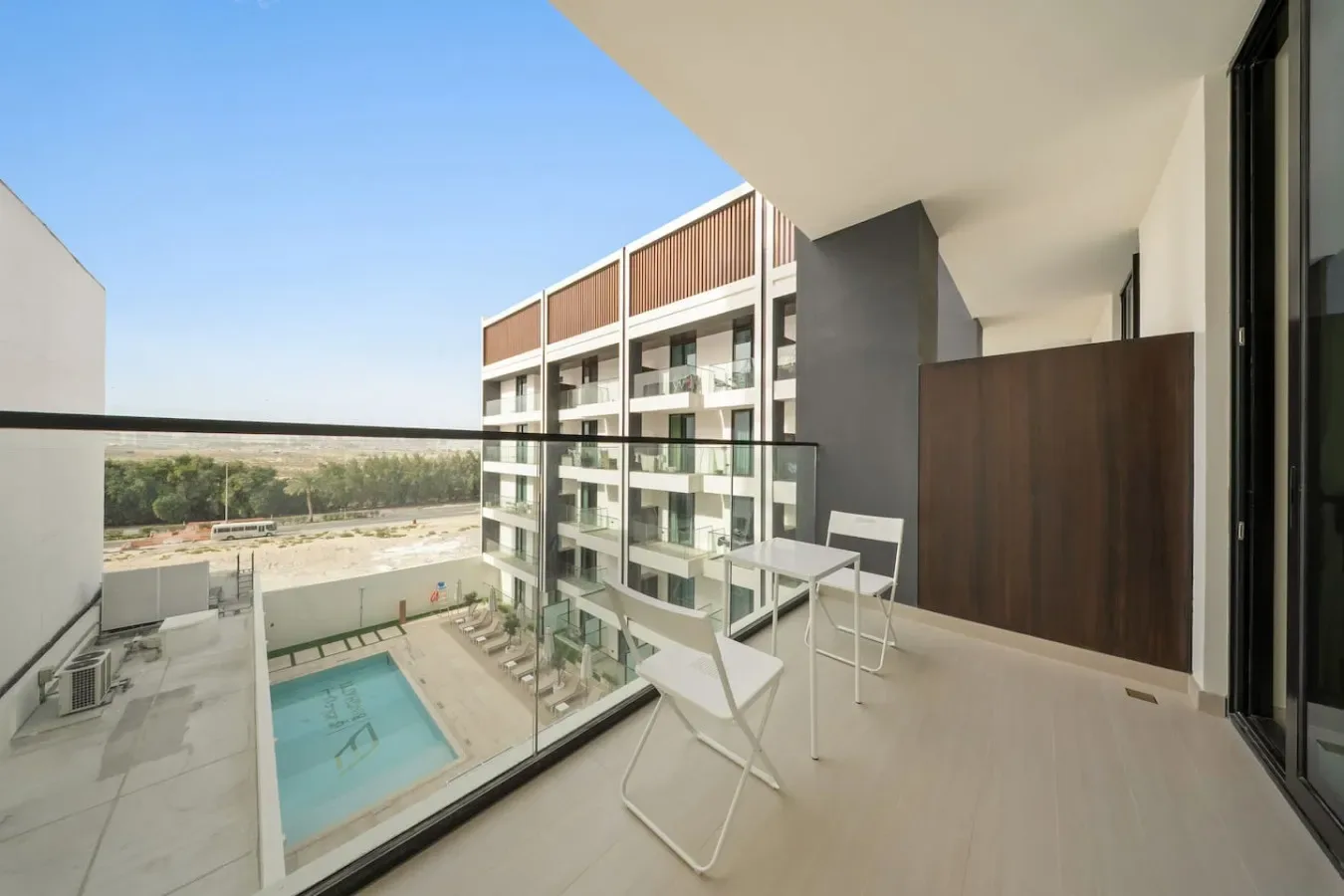 Un balcón con barandilla de vidrio da a una piscina y edificios cercanos.