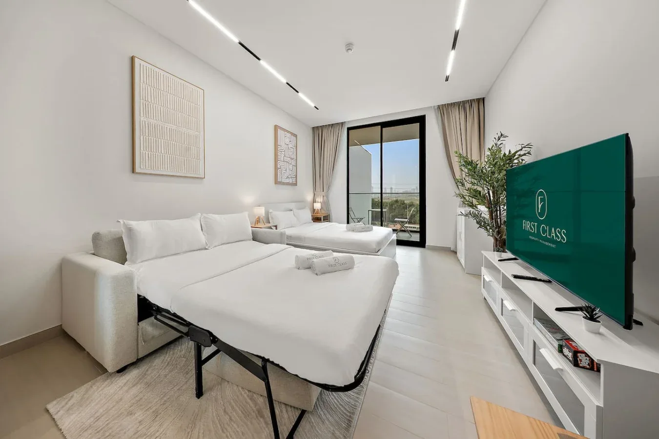 Un dormitorio luminoso con camas blancas, un televisor y una puerta de cristal al balcón.