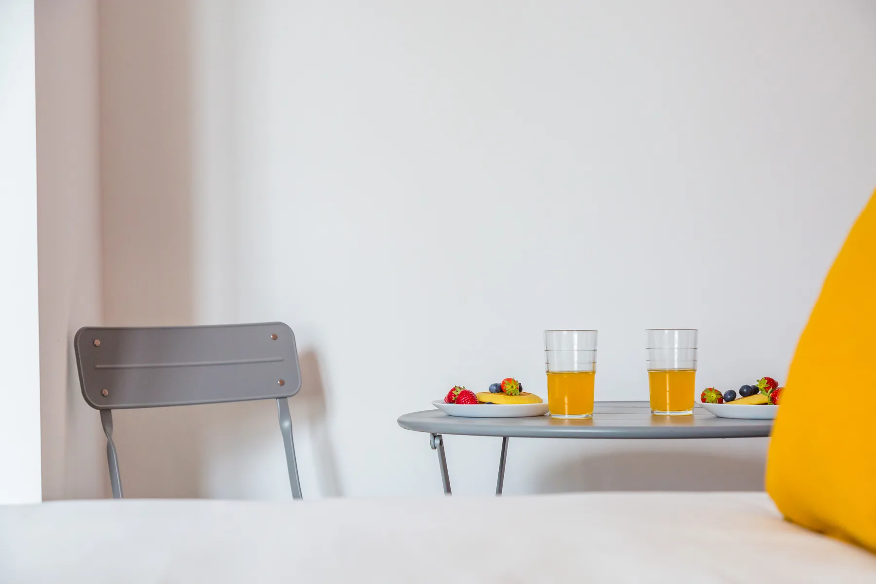Una mesa pequeña con bebidas y fruta está junto a una silla en una habitación luminosa.