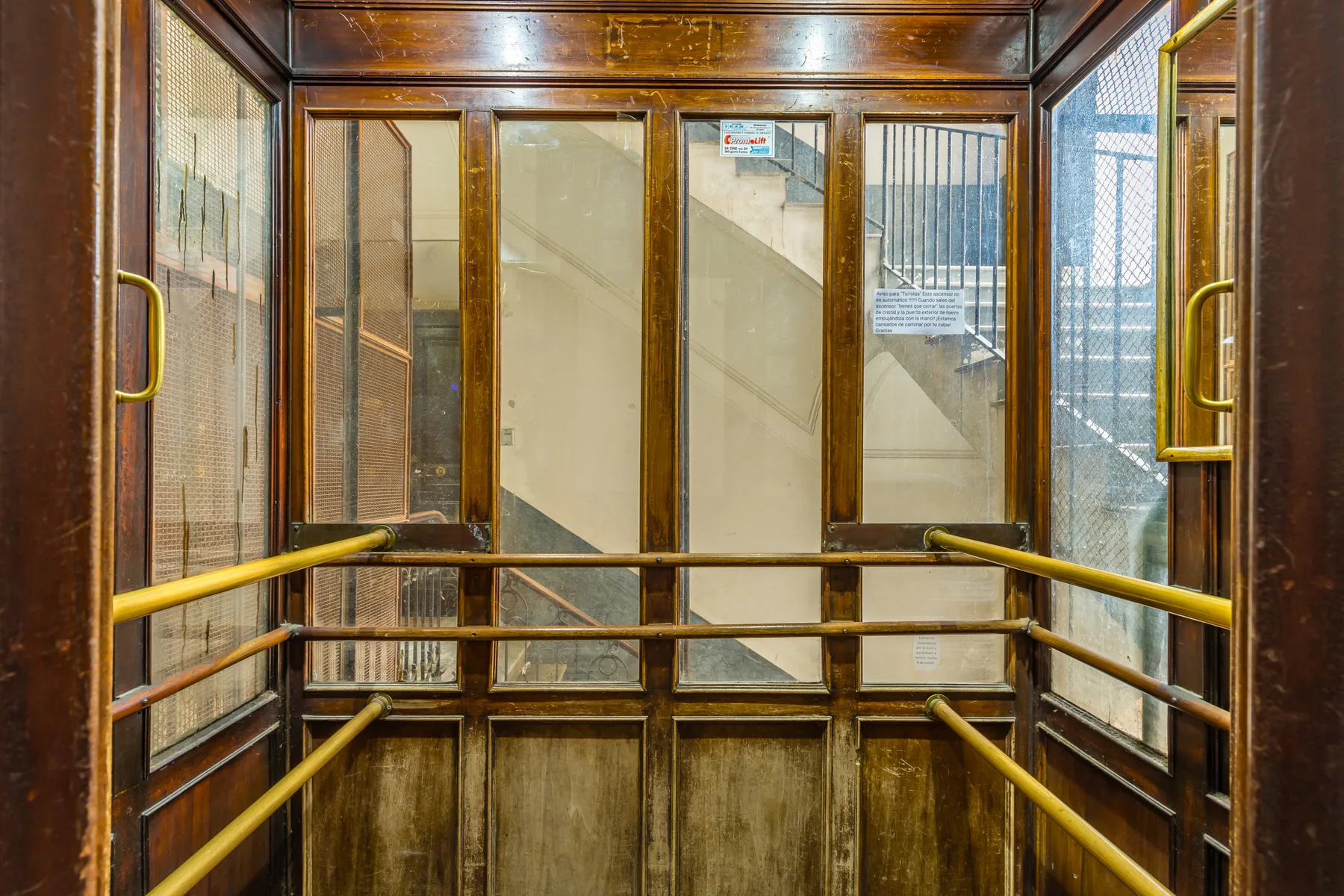 Interior de un ascensor revestido de madera, con paneles de vidrio y pasamanos metálicos.