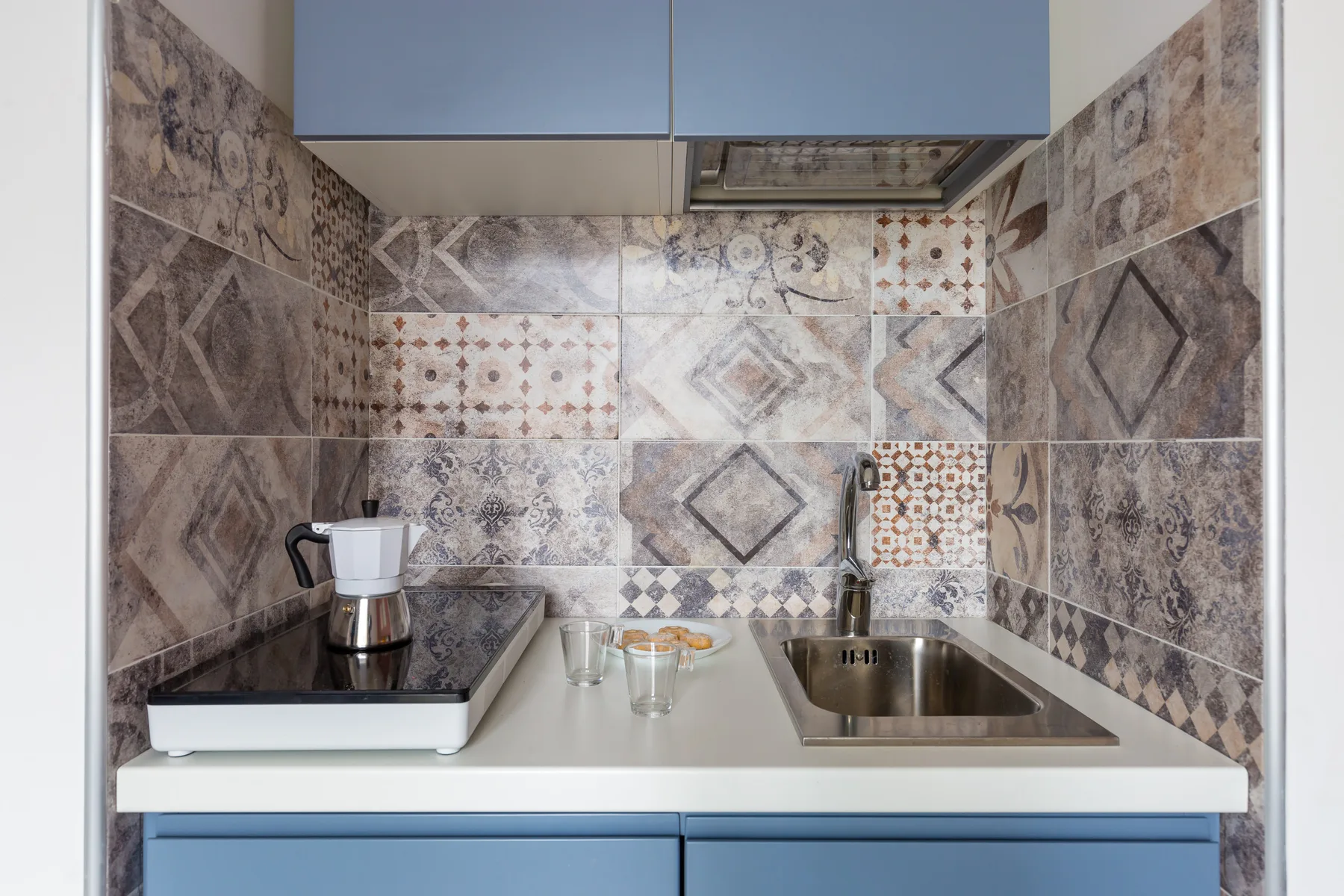 Una pequeña encimera de cocina con placa de cocción, fregadero y azulejos estampados.